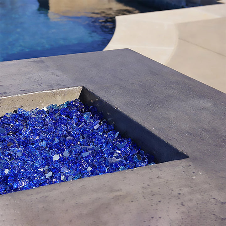 Close-up of the Prism Hardscapes Tavola 1 Gas Fire Pit in natural gray, showcasing vibrant blue fire glass. The fire pit's square design and intricate fire glass reflect a modern and stylish outdoor fire feature, ideal for creating ambiance in any outdoor space. This detailed view highlights the quality craftsmanship and aesthetic appeal of the fire pit.