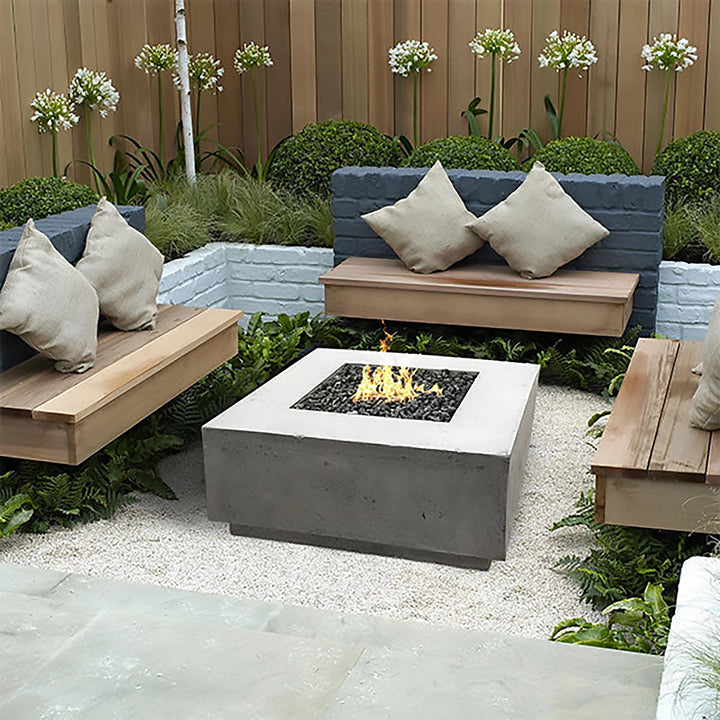 A beautifully designed Prism Hardscapes Tavola 2 Gas Fire Pit in natural gray, perfectly set in a serene garden patio with lush greenery and wooden seating. The modern square concrete fire pit enhances the outdoor living space with its clean lines and elegant fire display, ideal for backyard gatherings. This outdoor gas fire pit is a luxurious addition to any garden, adding warmth and style to your patio.