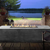 The Prism Hardscapes Tavola 4 Gas Fire Pit illuminates an evening patio setting, casting a warm glow over a scenic lakeside view. Its rectangular design and concrete finish provide a sophisticated touch to the patio space, making it the perfect addition for evening relaxation and outdoor entertainment. Ideal for those who enjoy the outdoors, this gas fire pit is both stylish and functional. 