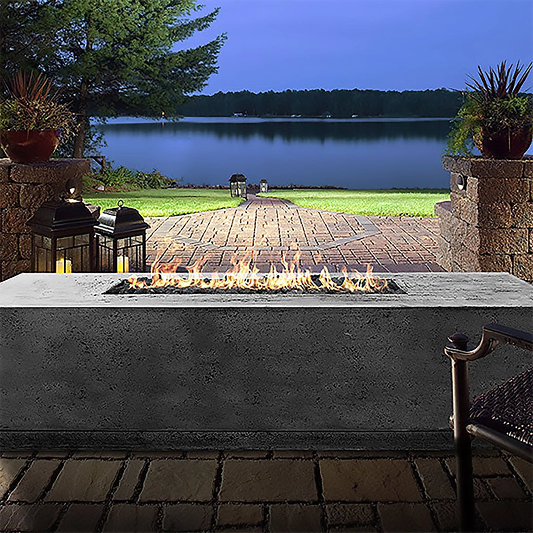 The Prism Hardscapes Tavola 4 Gas Fire Pit illuminates an evening patio setting, casting a warm glow over a scenic lakeside view. Its rectangular design and concrete finish provide a sophisticated touch to the patio space, making it the perfect addition for evening relaxation and outdoor entertainment. Ideal for those who enjoy the outdoors, this gas fire pit is both stylish and functional. 