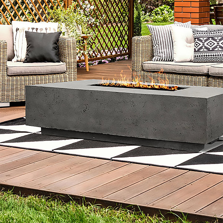 The Prism Hardscapes Tavola 4 Gas Fire Pit is featured in a cozy garden lounge, surrounded by wicker chairs and colorful cushions. The rectangular fire pit, with its sturdy concrete design, complements the natural garden setting while providing a warm and inviting flame. This gas fire pit is ideal for backyard gatherings, creating a focal point for outdoor entertainment. 