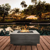 The Prism Hardscapes Tavola 42 Gas Fire Pit in a contemporary evening poolside setting with soft, ambient lighting. The natural gray concrete finish is elegantly illuminated by the bright, warm flames, reflecting in the pool. Its clean, geometric design enhances the modern outdoor aesthetic, perfect for hosting evening events or intimate gatherings. This luxury fire pit serves as a striking focal point in sophisticated backyards or poolside areas. 