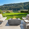 A scenic view of the Prism Hardscapes Tavola 42 Gas Fire Pit set on a spacious riverside patio. The sleek, modern design in a natural gray finish seamlessly blends with the surrounding nature, offering a perfect centerpiece for outdoor gatherings. Featuring a large burner that emits a bright flame, this contemporary fire pit enhances outdoor living spaces with its durable concrete construction. Ideal for backyards and patios, it offers style and function with clean, straight lines.