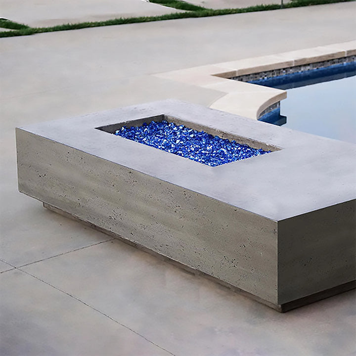 The Prism Hardscapes Tavola 5 Gas Fire Pit by a poolside setting, featuring a rectangular design with blue fire glass, adding a striking color contrast. This fire pit is ideal for contemporary outdoor spaces, offering warmth and ambiance near a pool. The smooth, concrete texture of the fire pit enhances the modern pool deck’s clean aesthetic.