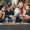 A group of friends enjoying an evening gathering around the Prism Hardscapes Tavola 5 Gas Fire Pit. The fire pit’s rectangular design in a sleek concrete finish offers a stylish centerpiece for social events. Its ample burner area provides a warm, inviting flame for a cozy outdoor atmosphere. Perfect for casual dining or relaxed conversations in the backyard. 