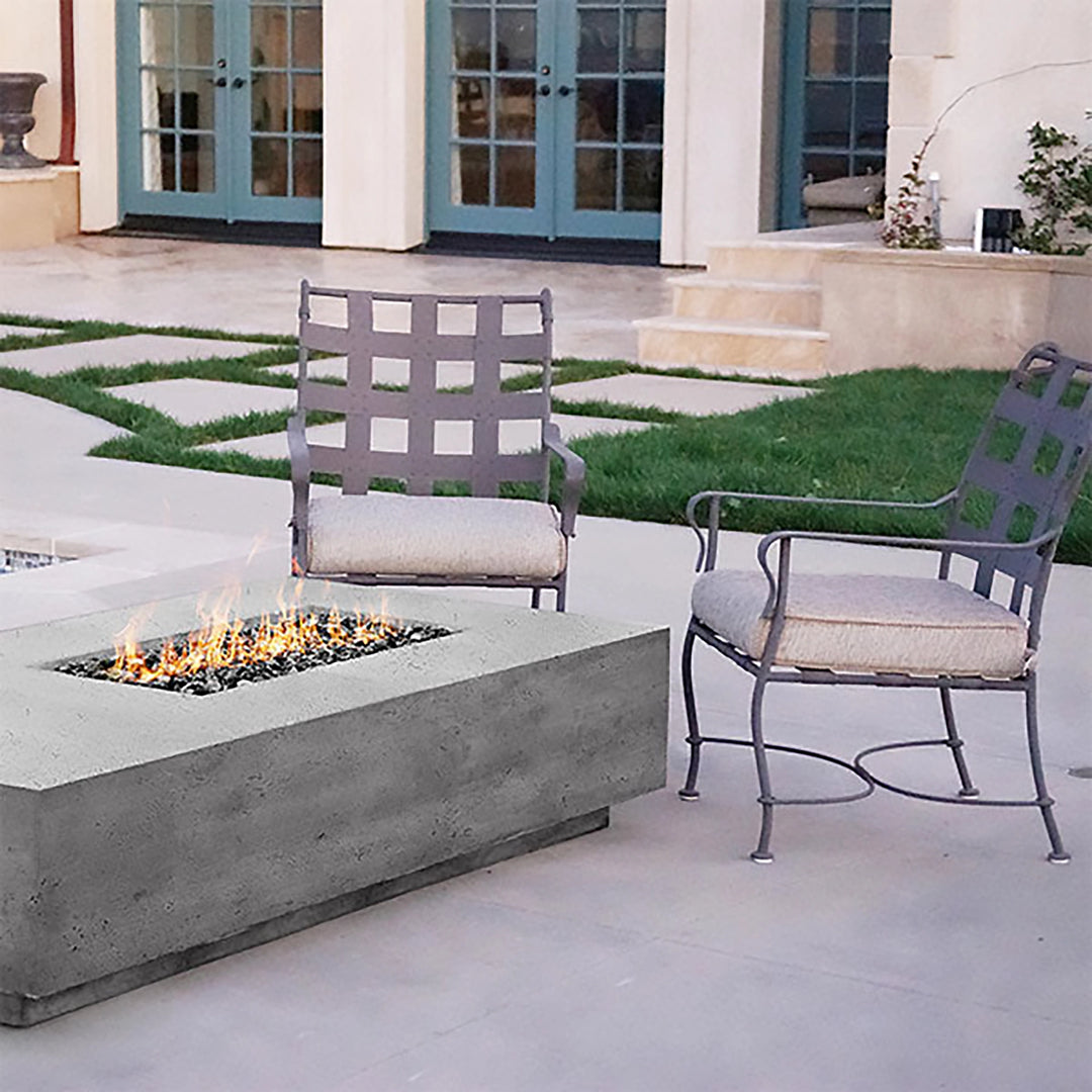 A modern outdoor patio featuring the Prism Hardscapes Tavola 5 Gas Fire Pit in a smooth concrete finish. The fire pit is accompanied by metal-framed chairs with neutral cushions, creating a cozy setting for outdoor relaxation. The rectangular fire pit’s sleek design complements the surrounding architecture, making it a perfect addition to any luxury backyard space. 