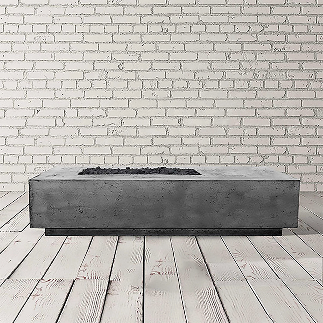 Prism Hardscapes Tavola 5 gas fire pit in pewter, displayed in a stylish studio with a white brick background. This sleek, rectangular fire pit has a medium-gray concrete finish, perfect for modern outdoor areas, featuring a recessed burner area filled with black lava rocks. 