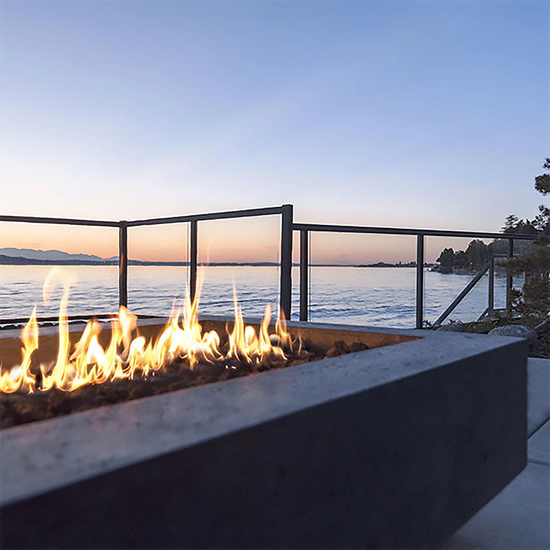 A serene lakeside evening with a lit Prism Hardscapes Tavola 6 Gas Fire Pit in the foreground. The fire pit's concrete structure adds to the tranquil setting, providing warmth as the sun sets over the water. Ideal for waterfront patios or decks, this fire pit offers a perfect blend of modern style and natural beauty.