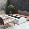  modern patio setup featuring the Prism Hardscapes Tavola 6 Gas Fire Pit, flanked by stylish woven chairs. The concrete fire pit blends seamlessly with the sleek, contemporary decor, offering both warmth and sophistication for outdoor entertaining. Ideal for residential or commercial patios.