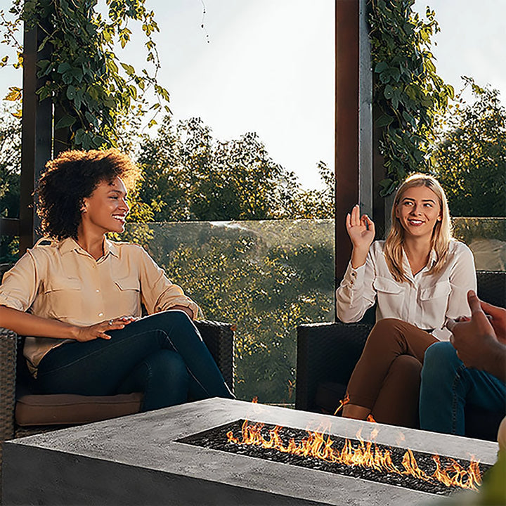 Two women enjoying a cozy evening outdoors, sitting next to a Prism Hardscapes Tavola 6 Gas Fire Pit, with flames dancing in the center. The fire pit is positioned in a modern outdoor setting, providing a warm and welcoming ambiance. This contemporary concrete fire pit is perfect for outdoor patios or terraces. 