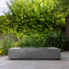 Outdoor lifestyle setting showcasing the Prism Hardscapes Tavola 72 Gas Fire Pit in gray finish. Positioned in a lush green garden, this sleek, modern fire pit blends perfectly with the natural surroundings, providing a stylish focal point for outdoor spaces. Ideal for contemporary patios and backyards, this rectangular fire pit combines modern design with high functionality, creating a cozy and inviting atmosphere for gatherings. Perfect for outdoor entertaining, luxury patios, and modern garden settings