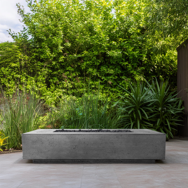 Outdoor lifestyle setting showcasing the Prism Hardscapes Tavola 72 Gas Fire Pit in gray finish. Positioned in a lush green garden, this sleek, modern fire pit blends perfectly with the natural surroundings, providing a stylish focal point for outdoor spaces. Ideal for contemporary patios and backyards, this rectangular fire pit combines modern design with high functionality, creating a cozy and inviting atmosphere for gatherings. Perfect for outdoor entertaining, luxury patios, and modern garden settings