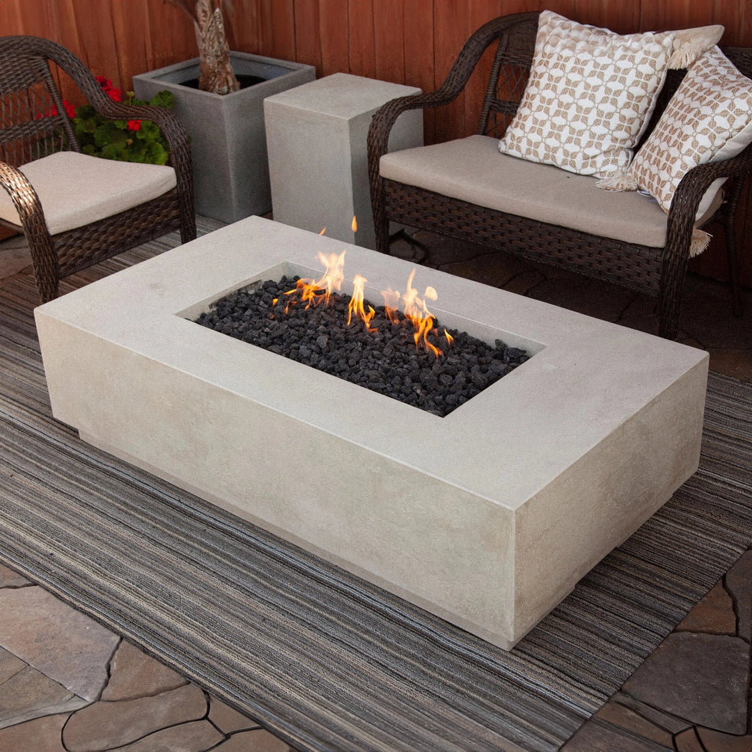 Prism Hardscapes Tavola 8 Gas Fire Pit in a natural finish showcased in an elegant outdoor patio setting. The rectangular fire pit is constructed from durable concrete with a smooth natural gray surface, complemented by lava rocks in the burner. Positioned on a striped outdoor rug, the fire pit provides a modern yet minimalist design, perfect for creating a warm and inviting space. Ideal for outdoor lounging, this gas fire pit combines contemporary aesthetics with functionality.