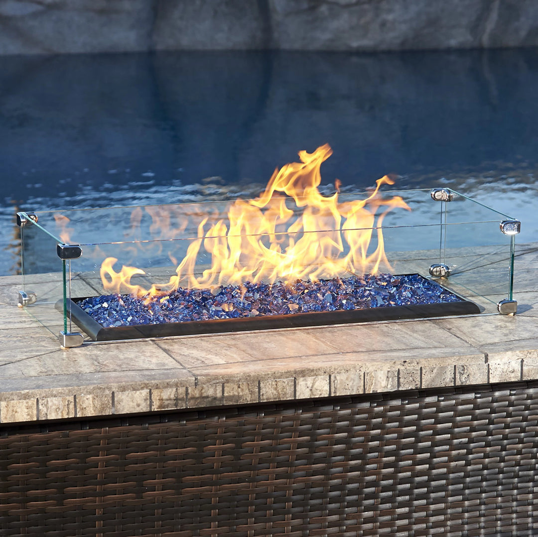 The American Fireglass Rectangular Glass Flame Guard is elegantly displayed on a wicker-style outdoor table, positioned by the poolside. The clear glass guard enhances flame stability while protecting the fire from wind, ensuring a vibrant and consistent flame over beautiful blue fire glass. This flame guard is a perfect accessory for outdoor fire pits, adding both safety and modern style to any poolside or patio fire table setup.