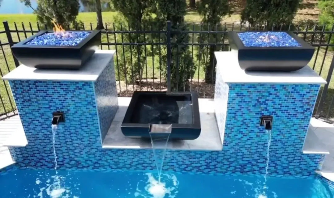 A water bowl pours into a pool in between two pillars with fire bowls on top in a backyard setting.