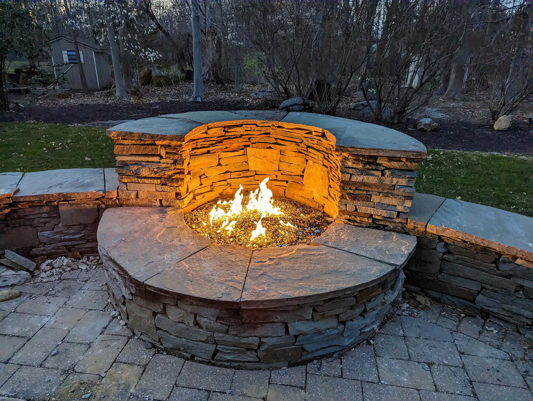 A custom fire pit  inlaid with slabs of stone showcases clear starfire glass overlaying a 24 burner ring ignited at dusk