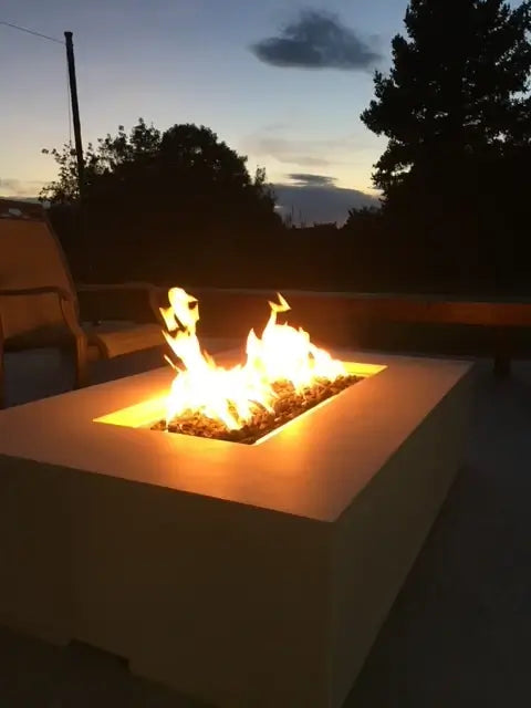 A closeup of a white concrete gas fire pit illuminated at dus on a beautiful forest patio.