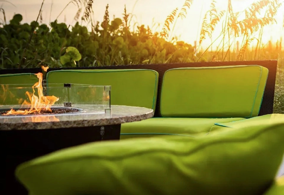 Elegant round fire pit with granite top and glass wind guard, set in a cozy outdoor lounge with vibrant green cushions at sunset.