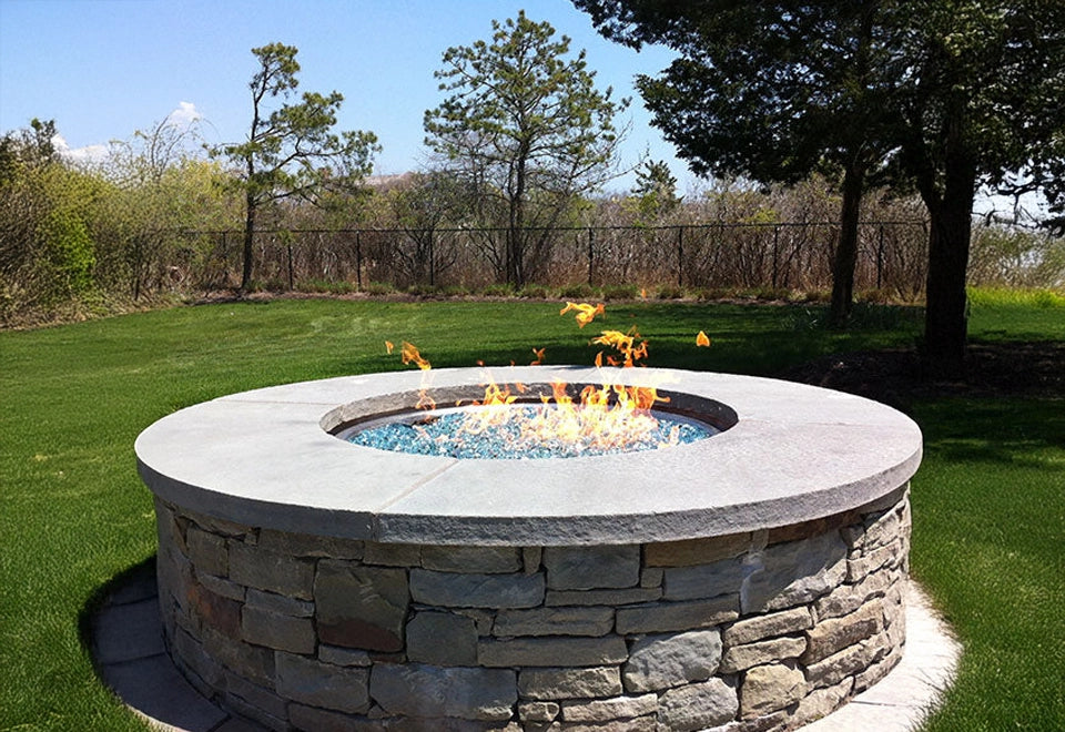 Traditional round stone gas fire pit with a smooth concrete top and vibrant flames over blue fire glass media, featuring a reliable electronic ignition system. Perfect for backyard fire pit installations, combining rustic charm with modern convenience for outdoor gatherings and landscaping projects.