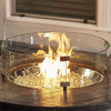 A 25-inch round tempered glass wind guard placed on a wooden table surrounding a fire pit, glowing with a vibrant flame and filled with fire glass. This elegant glass wind guard adds safety and style to the fire feature, making it a perfect addition to any outdoor patio or deck setup. The wind guard ensures stable flames while providing a clear, unobstructed view. 