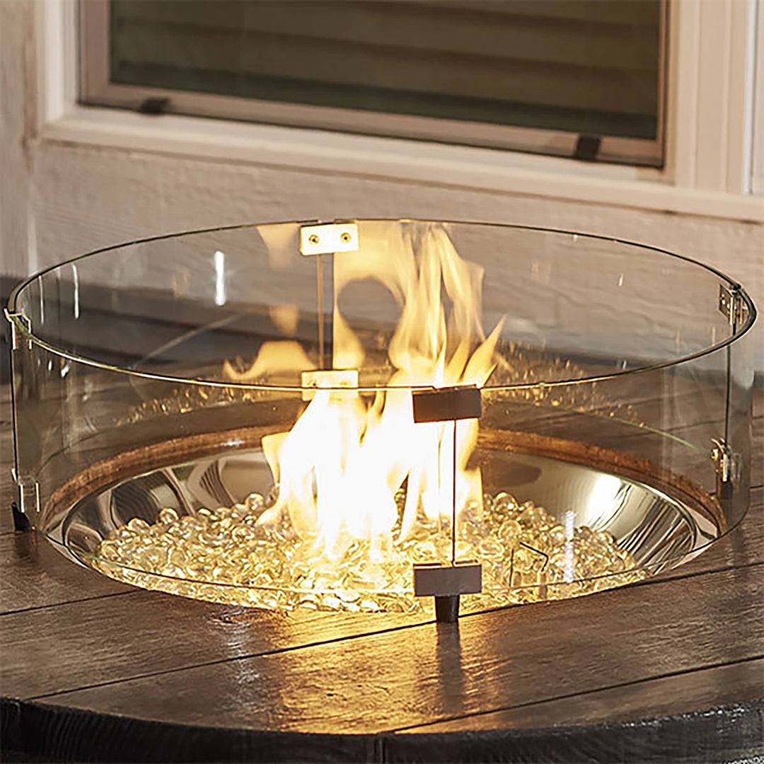 A 25-inch round tempered glass wind guard placed on a wooden table surrounding a fire pit, glowing with a vibrant flame and filled with fire glass. This elegant glass wind guard adds safety and style to the fire feature, making it a perfect addition to any outdoor patio or deck setup. The wind guard ensures stable flames while providing a clear, unobstructed view. 