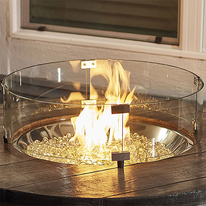 A 25-inch round tempered glass wind guard placed on a wooden table surrounding a fire pit, glowing with a vibrant flame and filled with fire glass. This elegant glass wind guard adds safety and style to the fire feature, making it a perfect addition to any outdoor patio or deck setup. The wind guard ensures stable flames while providing a clear, unobstructed view. 