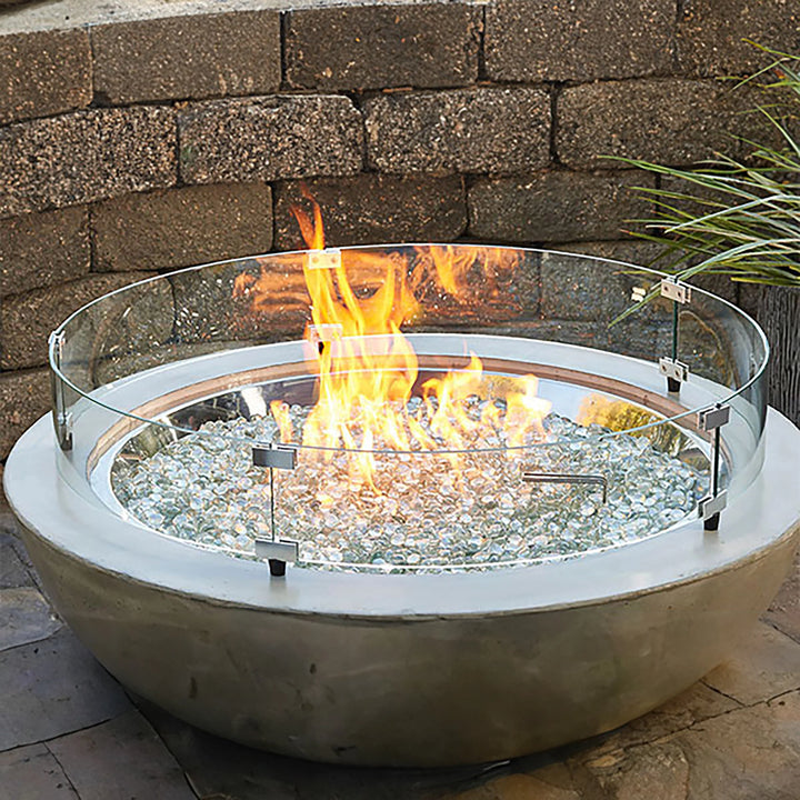A round 30-inch tempered glass wind guard surrounding an outdoor fire pit, filled with clear fire glass. The glass wind guard helps to protect the flames from the wind while providing an elegant, transparent barrier. Perfect for outdoor patios, this fire pit setup enhances the ambiance with the warm glow of fire against the backdrop of rustic stone.