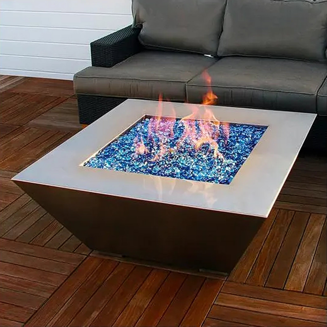 Modern square-shaped stainless steel fire pit filled with blue fire glass, set on a wooden deck beside a dark wicker outdoor sofa with gray cushions.