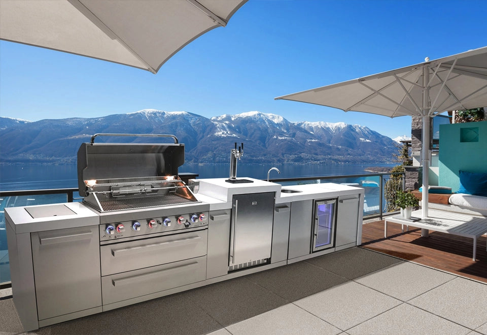 Modern stainless steel outdoor kitchen island with built-in grill, sink, refrigerator, and beverage cooler overlooking a stunning mountain lake view.