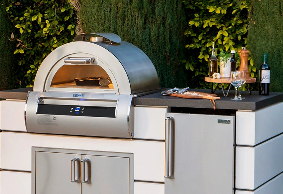 Modern stainless steel outdoor pizza oven with digital controls, built into a stylish white and black countertop kitchen setup, surrounded by fresh herbs and wine