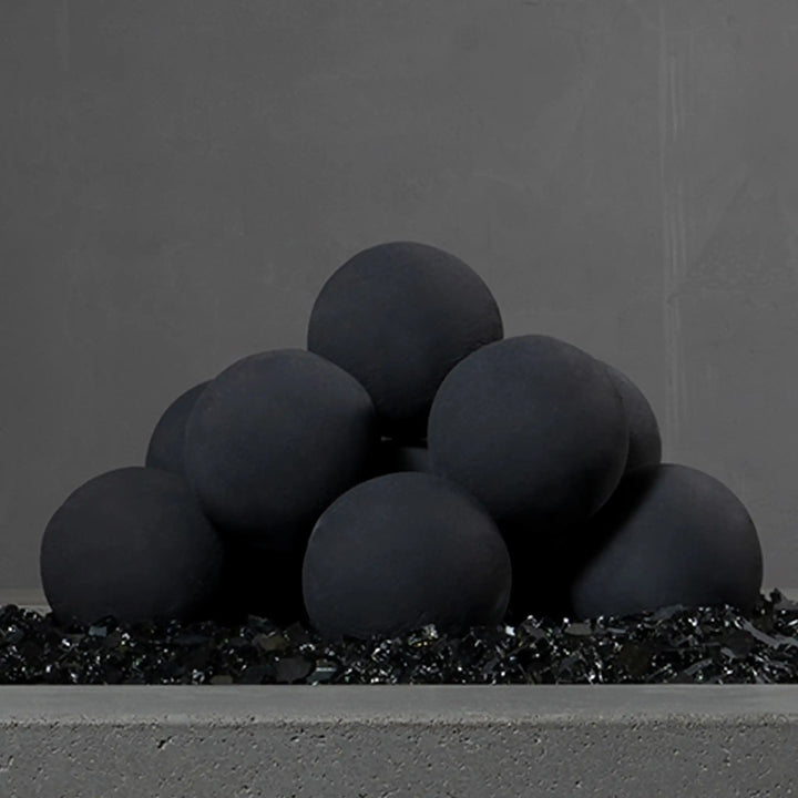 Close-up view of Starfire Designs 4-inch ceramic fire balls set of 12 in dark gray, stacked in a pyramid on a bed of black fire glass in an unlit fire feature. Their smooth texture and modern style enhance any contemporary fire pit setup