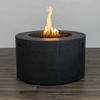 In a simple studio setting, a tall dark gray round fire pit stands on a wood-grain floor, its cylindrical concrete fire pit body showing slim handholds on each side and a recessed opening filled with small brown stones, where a vertical flame rises from the center of the gas fire pit, the plain background and even lighting emphasizing the smooth surface and proportions that give this piece the look of a straightforward modern fire pit.