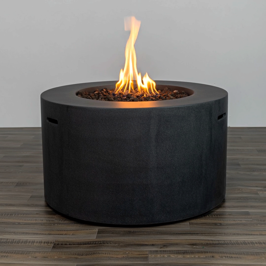 In a simple studio setting, a tall dark gray round fire pit stands on a wood-grain floor, its cylindrical concrete fire pit body showing slim handholds on each side and a recessed opening filled with small brown stones, where a vertical flame rises from the center of the gas fire pit, the plain background and even lighting emphasizing the smooth surface and proportions that give this piece the look of a straightforward modern fire pit.