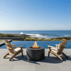 On a pale stone terrace beside an infinity pool that blends into a bright blue ocean, a dark gray round fire pit burns between two light wood Adirondack chairs with cream cushions, the low outdoor fire pit positioned close to the water while grasses border the pool and waves crash on distant rocks, forming a simple patio fire pit arrangement that turns the open deck into a calm backyard oasis under a clear midday sky.