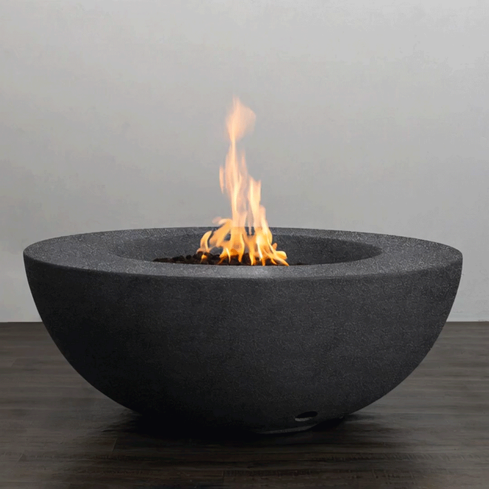 A low, graphite-colored round fire pit with a smooth, sculptural bowl shape sends a steady column of flame upward from a bed of dark lava rocks, the minimalist concrete fire pit resting on a wood-look patio surface and showcasing a clean-lined modern fire pit design, powered by a hidden natural gas fire pit burner that delivers consistent heat and an elegant focal point for contemporary outdoor lounges and compact urban terraces.