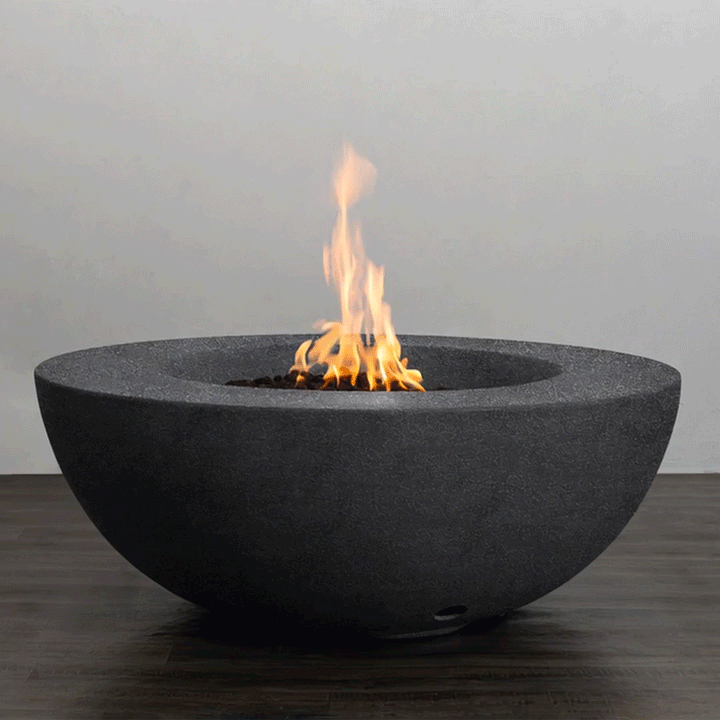 A low, graphite-colored round fire pit with a smooth, sculptural bowl shape sends a steady column of flame upward from a bed of dark lava rocks, the minimalist concrete fire pit resting on a wood-look patio surface and showcasing a clean-lined modern fire pit design, powered by a hidden natural gas fire pit burner that delivers consistent heat and an elegant focal point for contemporary outdoor lounges and compact urban terraces.