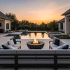A spacious backyard with a shimmering pool and manicured landscaping features a dark graphite round concrete fire pit centered before an L-shaped outdoor sofa, its bright flame flickering above lava rocks as the sky glows at dusk; this stylish backyard fire pit becomes the heart of the patio, ideal for outdoor entertaining with friends, offering a modern yet inviting place to curl up with blankets, cocktails, and conversation long after the sun goes down.