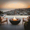 On an elevated terrace overlooking a coastal city and calm ocean at twilight, a sleek graphite round fire pit blazes between two cushioned wood lounge chairs, the flames rising cleanly from a recessed burner within the bowl; this streamlined outdoor fire pit, fueled by a convenient natural gas fire pit system, acts as a modern fire pit centerpiece for the balcony, inviting quiet moments, warm drinks, and panoramic views as city lights start to twinkle below.