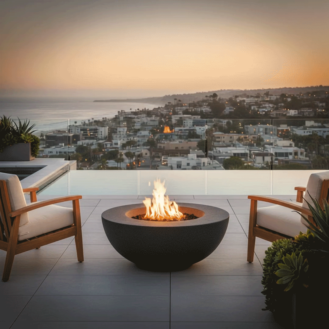 On an elevated terrace overlooking a coastal city and calm ocean at twilight, a sleek graphite round fire pit blazes between two cushioned wood lounge chairs, the flames rising cleanly from a recessed burner within the bowl; this streamlined outdoor fire pit, fueled by a convenient natural gas fire pit system, acts as a modern fire pit centerpiece for the balcony, inviting quiet moments, warm drinks, and panoramic views as city lights start to twinkle below.