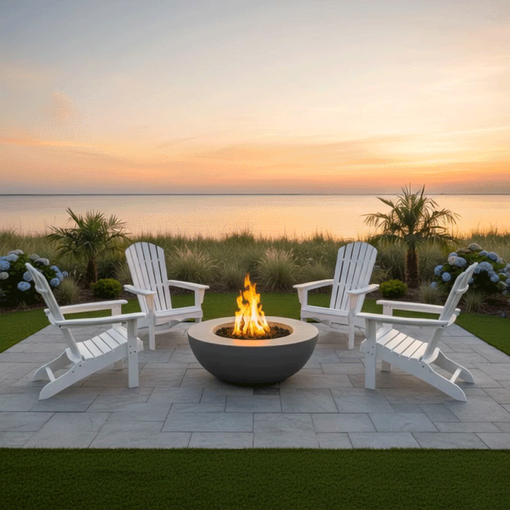 On a square stone patio set into a green lawn near calm water at sunset, a gray bowl-shaped backyard fire pit with a wide rim glows at the center of four white Adirondack chairs, the low form filled with dark lava rocks while ornamental grasses, small palms, and blue hydrangeas frame the edges of the seating area, the arrangement reading as a relaxed outdoor fire pit circle and quiet family gathering fire pit beside the soft orange and pink sky.