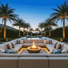 In a large sunken seating area framed by tall palms and hedges, a shallow concrete fire pit with a sand-colored bowl shape burns at the center of an expansive U-shaped sectional, the low flame flickering above dark stones while warm strip lighting traces the sofa base, and beyond the steps a long pool and rows of illuminated tree trunks stretch toward the horizon, forming a symmetrical fire feature clearly arranged for outdoor entertaining within a sheltered backyard oasis.