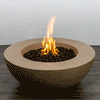 In a simple studio scene, a sand-colored concrete fire pit shaped like a wide, shallow fire bowl sits on a dark wood floor against a plain pale wall, its thick rounded rim dropping to a recessed basin filled with irregular black lava rocks, where a bright orange flame rises in loose vertical strands, the clean curves and unadorned surroundings emphasizing the low modern fire pit as the only object in the frame.