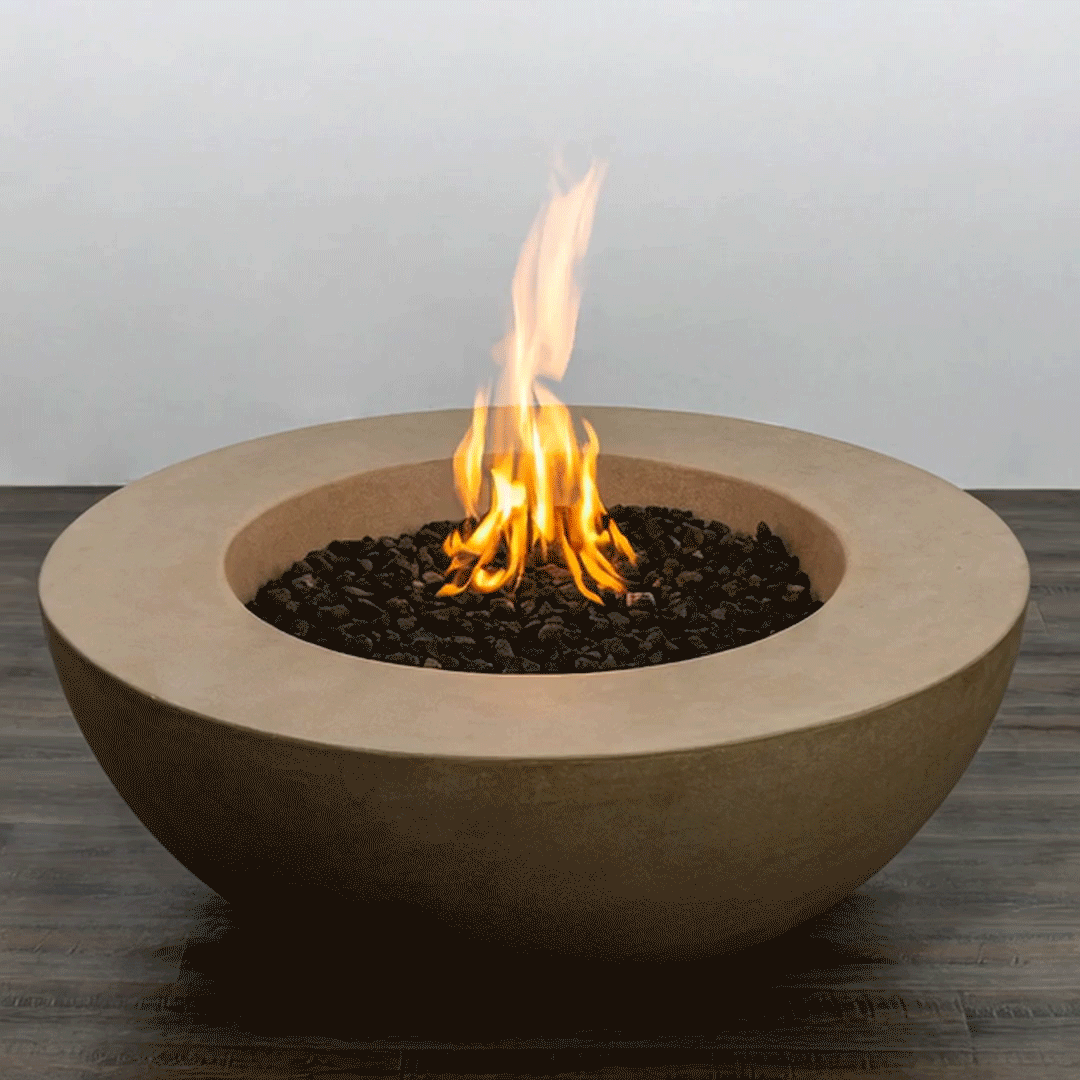 In a simple studio scene, a sand-colored concrete fire pit shaped like a wide, shallow fire bowl sits on a dark wood floor against a plain pale wall, its thick rounded rim dropping to a recessed basin filled with irregular black lava rocks, where a bright orange flame rises in loose vertical strands, the clean curves and unadorned surroundings emphasizing the low modern fire pit as the only object in the frame.