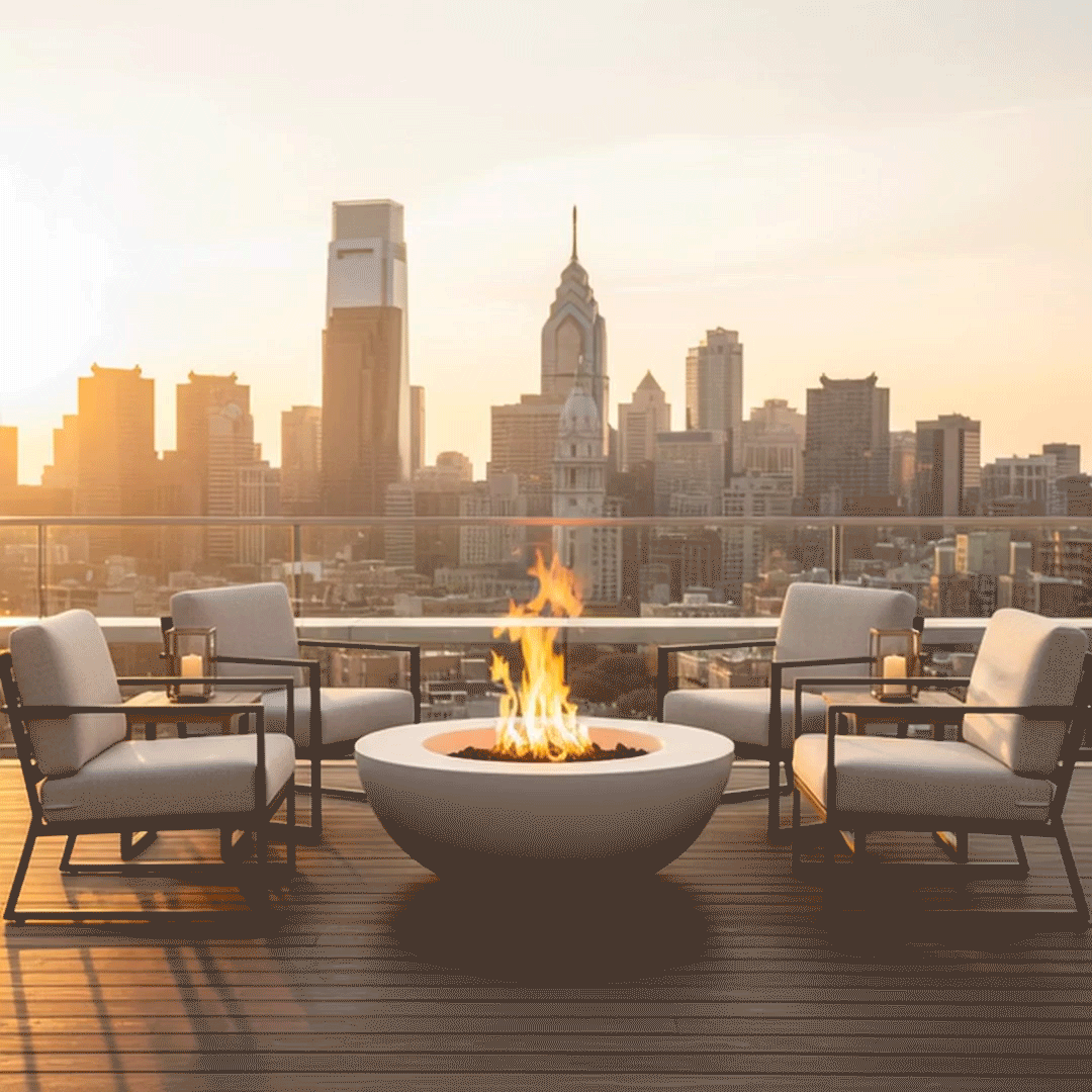 On a high city terrace in soft daylight, a white concrete fire pit with a wide rounded bowl sits in the center of four metal-framed lounge chairs with pale cushions, the low form resting on narrow deck boards and holding a ring of dark stones and lively orange flames, while a glass railing lines the edge and a row of skyscrapers fills the hazy skyline, the simple layout presenting a modest outdoor fire pit that matches the surrounding modern fire pit aesthetic.