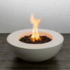 A low white concrete fire pit shaped like a wide, shallow fire bowl rests on a dark wood plank floor against a plain light wall, its thick rounded rim curving inward to a recessed center filled with dark porous lava rocks where a bright yellow flame rises in loose vertical strands, the simple geometry and smooth surface emphasizing a clean, modern fire pit with no chairs, plants, or other decor in view.