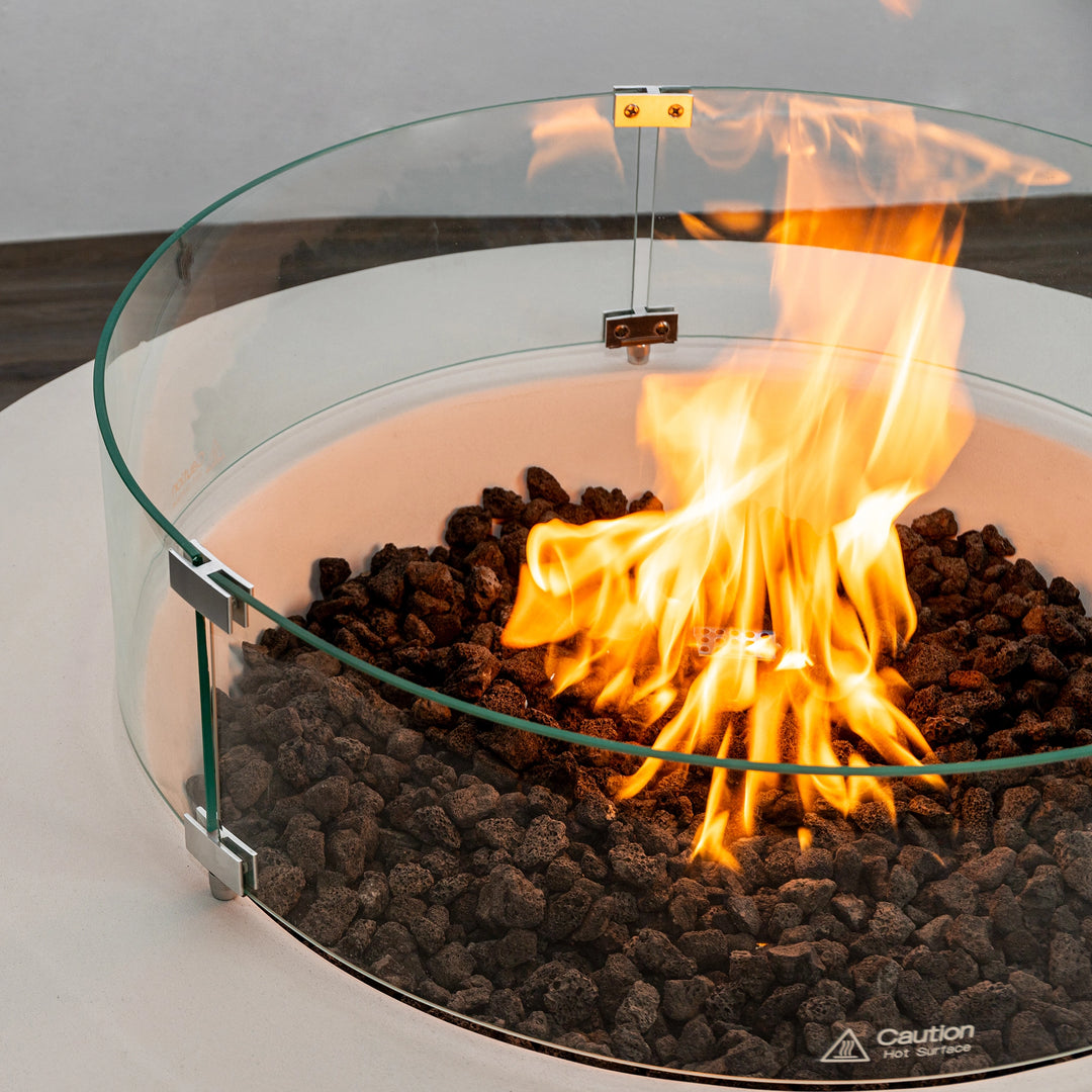 Close-up view of the Starfire Designs Beton 42 Inch White Round Concrete Gas Fire Pit with vibrant flames surrounded by black lava rocks and a glass wind guard, emphasizing its modern fire feature.