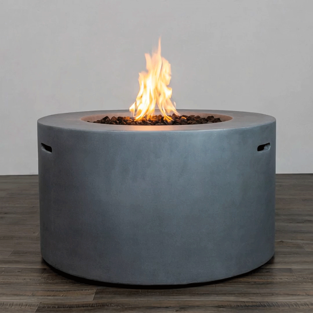 A cylindrical gray round fire pit sits on a wood-look floor against a plain backdrop, its thick concrete fire pit walls enclosing a recessed bed of lava rocks where a tall, steady flame burns from a hidden burner, emphasizing the clean, sculptural lines of this modern fire pit; the design suggests a low-maintenance gas fire pit ideal for contemporary patios, rooftop lounges, or courtyard seating areas that call for understated warmth and style.