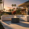 In a desert-inspired backyard with palm trees, cactus, and flowering shrubs, a dark gray concrete fire pit glows at the center of a large white sectional, the flames rising in a neat circle as sunset light washes over the tiled terrace; this inviting patio fire pit doubles as a sleek modern fire pit focal point, creating an effortless hub for outdoor entertaining where guests can lounge, share drinks, and enjoy warm evenings under an open sky.