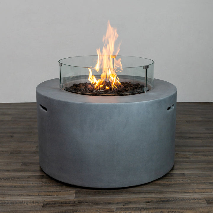 A side-angle view of the Starfire Designs Beton 42" Natural Gray Round Concrete Gas Fire Pit featuring a glass wind guard encircling the flame. The natural gray concrete body and glowing flame provide an elegant addition to any outdoor setting.