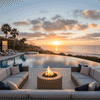 At sunset beside an infinity pool that seems to spill into the ocean horizon, a glowing round fire pit flickers between two deep, cushioned sectionals, casting warm light across the stone patio as palm trees and sailboats frame the view, creating an inviting outdoor fire pit scene that feels like a private resort, where this luxury fire pit anchors a serene backyard oasis designed for relaxed lounging, stargazing, and lingering conversations by the water’s edge.